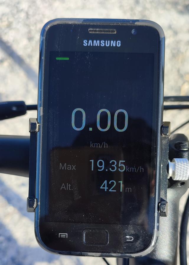 Photo of the device displaying the app's UI - white & gray text on a black background. The speed (currently 0.0) is large and centred, with the max speed and current altitude below it.